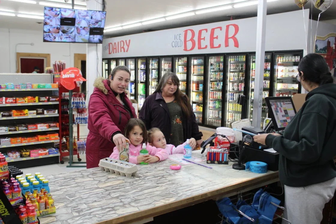 Convenience store checkout counter with cashier assisting customers near refrigerated beer and dairy product displays, typical of retail food distribution environments.